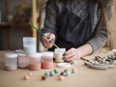 Hands painting with a brush, mixing colors in jars. Neutral background with craft beads on table. Calm, creative atmosphere.