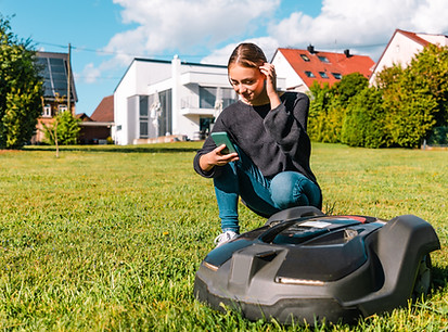Young woman controlling a robot lawn mower with her smart phone in the summer. The woman k