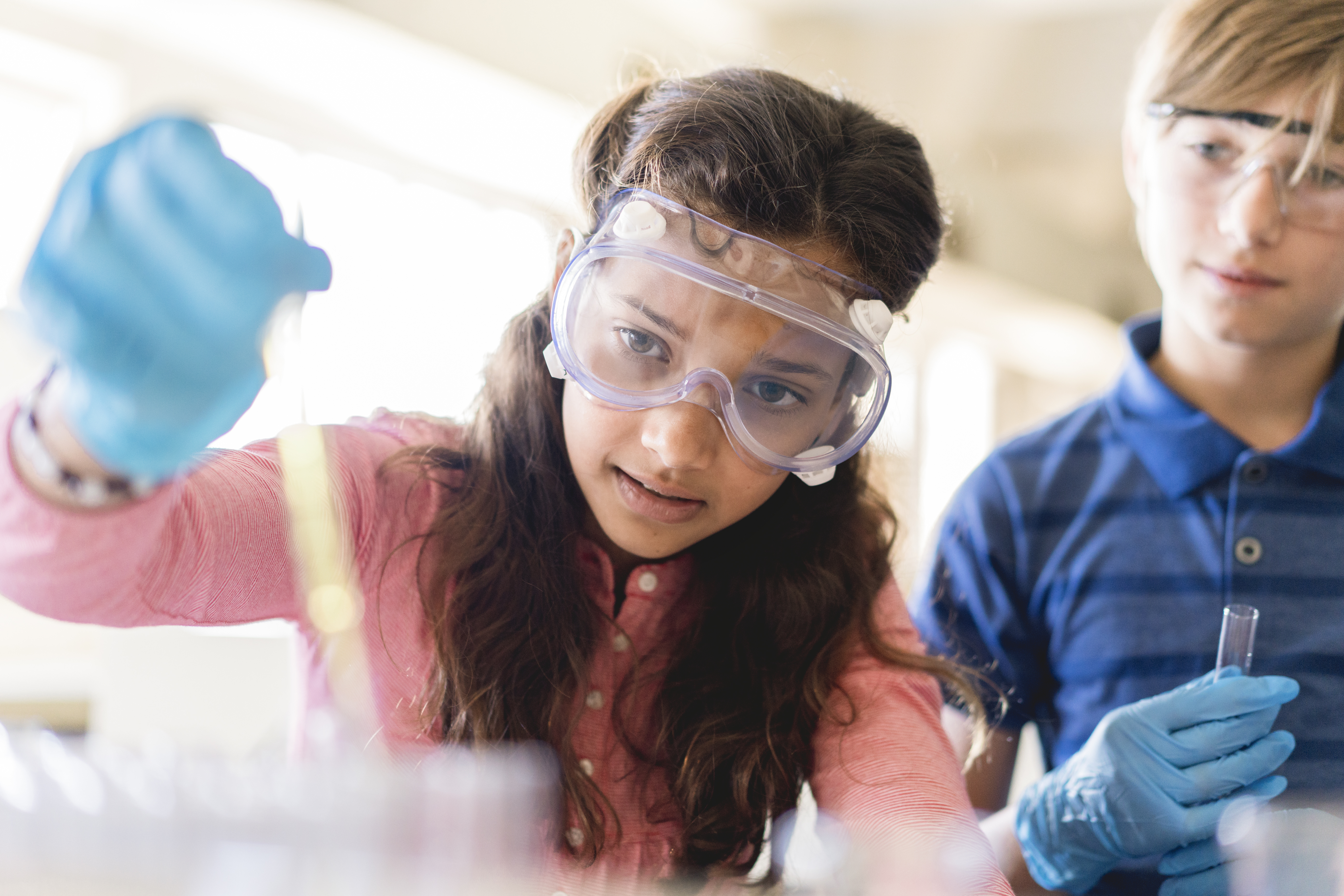 Two students in safety goggles and gloves conducting a science experiment with test tubes in a well-lit classroom.