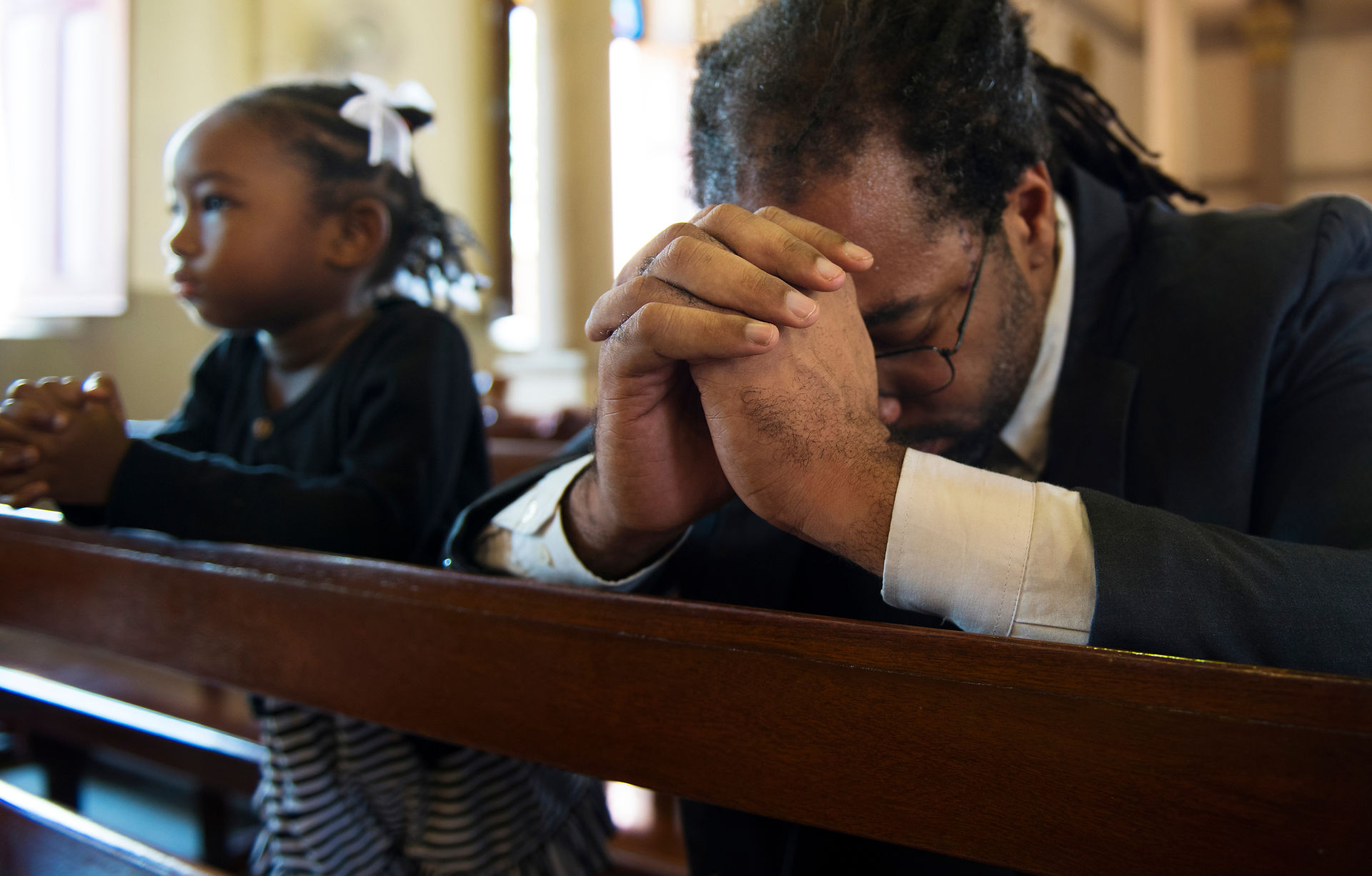 A young Black girl with braids in a black dress stands at a pew in prayer next to an older Black man with braids who has his head bowed to his clasped hands. 