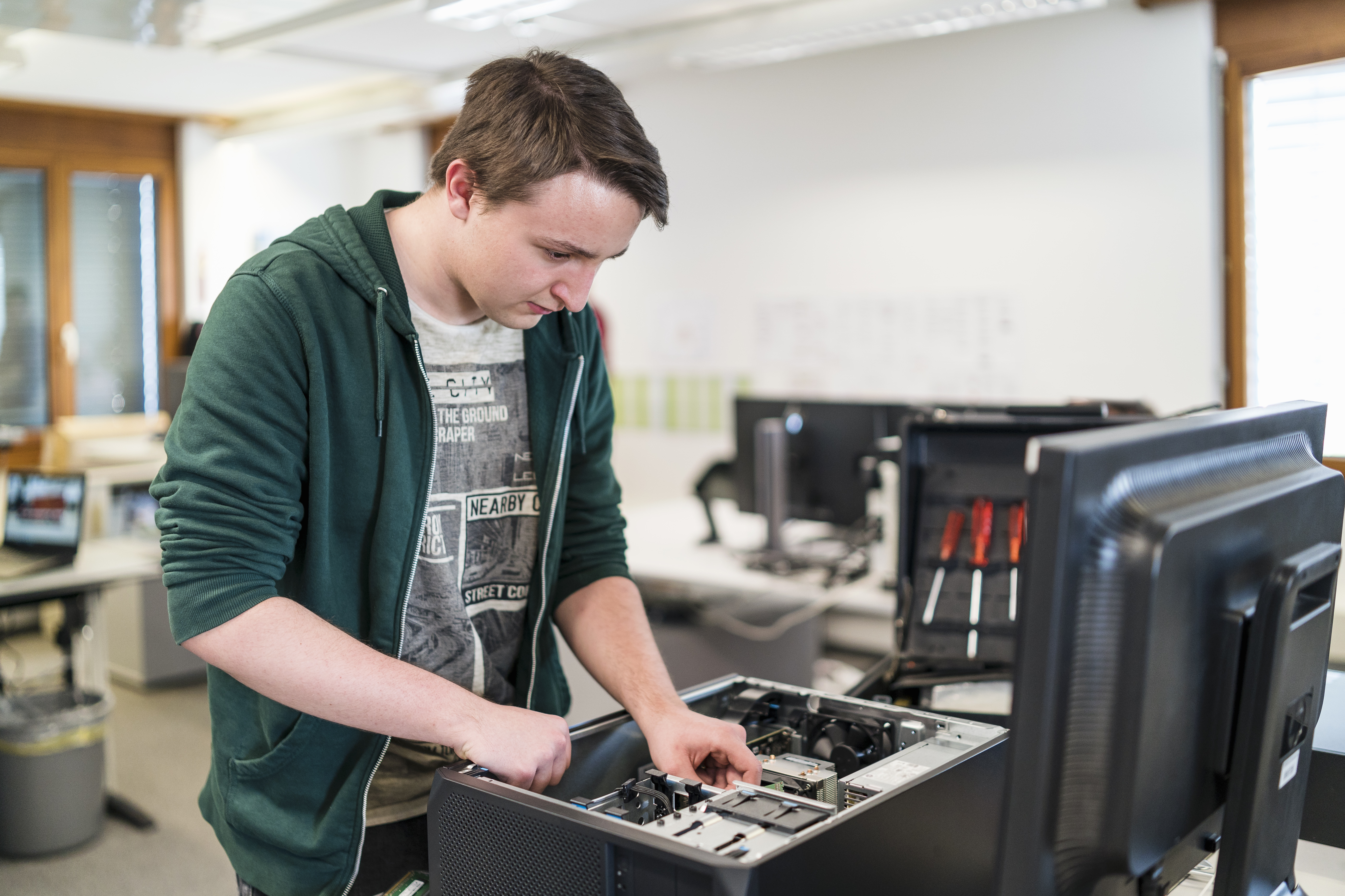 Young man in a green hoodie repairing or assembling a desktop computer inside a modern office. He is focused on working with
