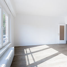 A bright, empty room with white walls, light wooden floors, and a window casting shadows