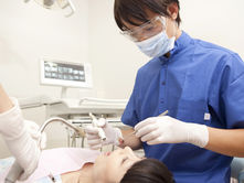 Dentist in blue scrubs examines a patient's mouth, using dental tools. Screen in background. Sterile, clinical environment.