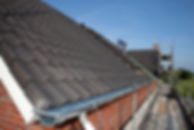 A worker installs dark tiles on a sloped roof, secured by scaffolding and safety gear