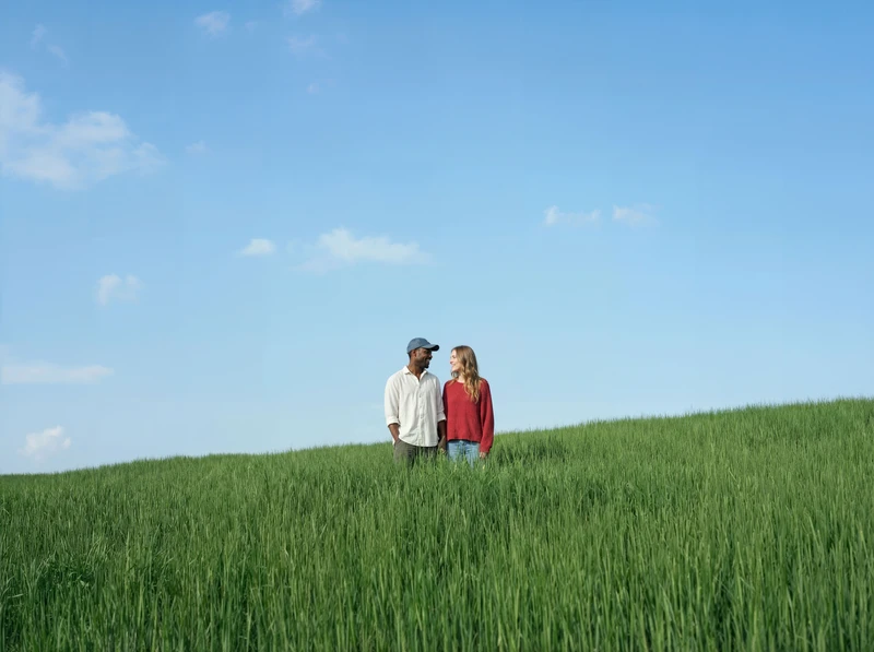 Ein Paar steht einander zugewandt in einem grünen Feld, im Hintergrund der blaue wolkenlose Himmel
