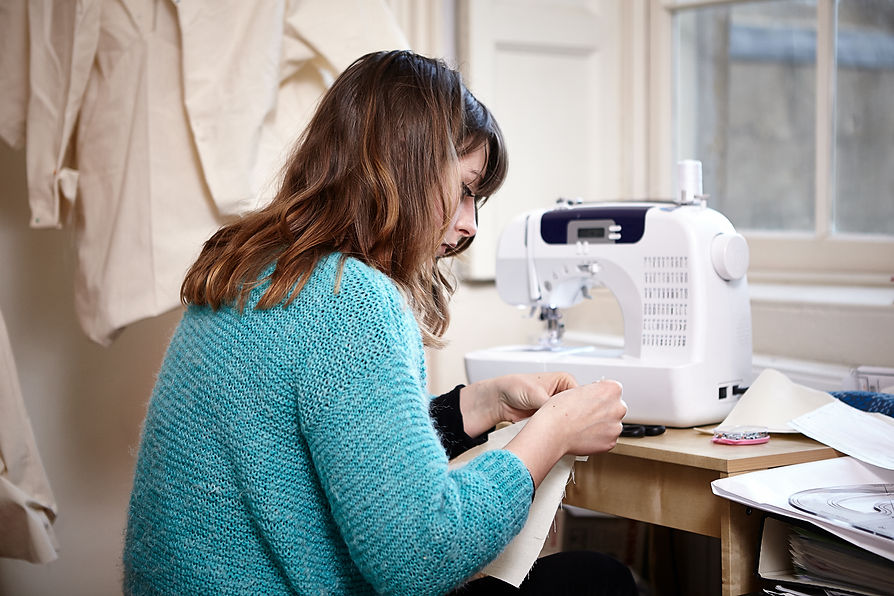 Young woman with brown hair in a teal sweater concentrating at a white sewing machine designing clothing
