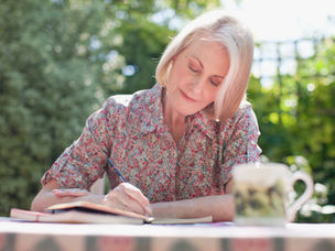 A woman in a floral shirt writes in a notebook at an outdoor table, next to a patterned mug; peaceful greenery in the background.