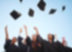 Graduates in black gowns cheerfully throw caps in the air against a clear blue sky, celebrating with joy and excitement.