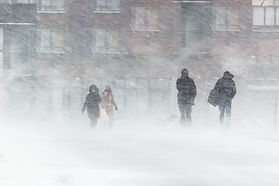 People walking in heavy snow with blurred buildings in the background. Winter coats and bags, grayish tones, stormy and cold mood.