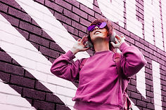 Young woman in pink sweatshirt and purple sunglasses adjusts headphones before a purple-and-white brick mural.