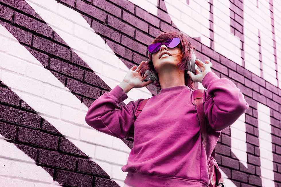 A woman with messy, side-swept reddish-pink hair stands in the bottom right corner, her body angled to the left against pink tinted brick wall with faint white graffiti. She wears a fuchsia sweatshirt, a dark pink backpack, and heart-shaped sunglasses while looking upward and holding a pair of over-ear headphones.