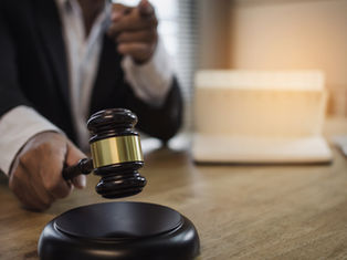 A person in a suit slams a gavel on a wooden desk, pointing forward. Books and papers are in the blurred background, lit warmly.