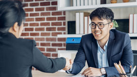 Two people in business attire shake hands in an office with a brick wall. One smiles warmly. Shelves with books and plants in the background.