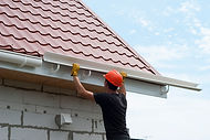 Worker in an orange hard hat and yellow work gloves installing a white plastic rain gutter onto a building beneath a red wavy tiled roof