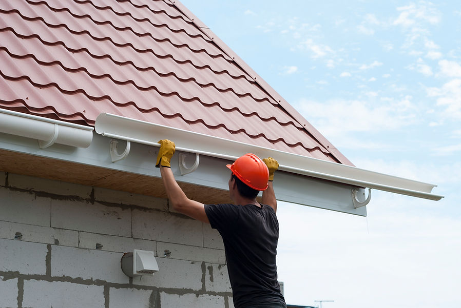 A roofer installing white gutters on a building with a red metal roof
