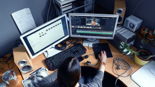 Person editing video on dual monitors at a desk with speakers, plants, and papers. The screens show a video timeline. Cozy workspace.