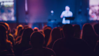 Blurred speaker on stage before a seated audience, bathed in blue and purple light at a live event