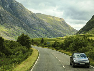 Car on a winding road through lush green mountains with cloudy skies. Rugged peaks and greenery create a serene, natural scene.