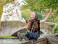 A smiling girl holds her arms up to the sky, sat on a wall alongside a river