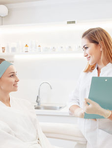 A woman in a headband and robe smiles at a nurse with a clipboard in a bright, white clinic. Both appear happy and engaged.