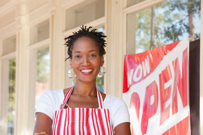 Energetic entrepreneur proudly standing by 'Now Open' sign, emphasizing the importance of solid financial planning in ensuring long-term business success and personal wealth management.