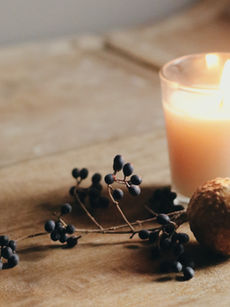Lit candle on a wooden table with dark berries and a textured brown sphere. Soft, warm lighting creates a cosy, serene atmosphere.