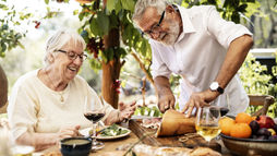 Smiling elderly couple shares an outdoor meal under leafy vines, with wine, fruit, and a man cutting bread on a wooden board.
