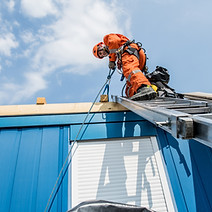 A worker in an orange suit and helmet is on a ladder, securing a rope on a blue building's roof