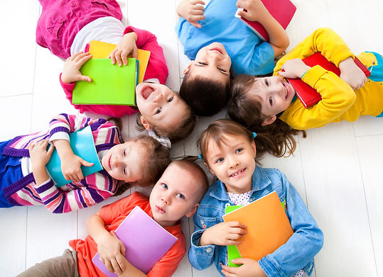 Six kids in colorful clothing lying in a circle on a white floor with their heads together and holding books