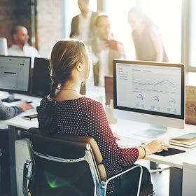 Femme et homme travaillant ensemble devant un ordinateur, analysant des graphiques financiers dans un environnement professionnel avec des collègues en arrière-plan
