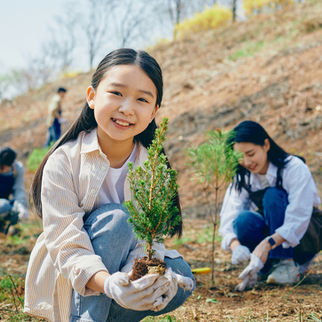 A young girl smiles proudly as she holds a sapling, participating in a community tree-planting event with others working in the background on a sunny day.