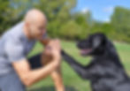 Veteran, mid-thirties waring shorts and a t-shirt gives a smiling black labrador retriever a high-fivel