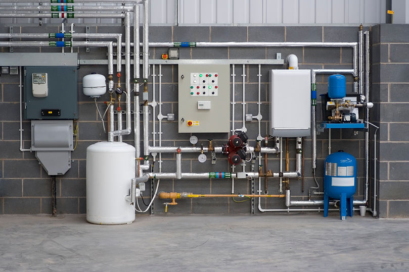 Industrial wall featuring complex grey pipework, electrical control boxes, and a blue expansion vessel