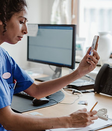 Nurse writing in clipboard while using phone for video conference at clinic