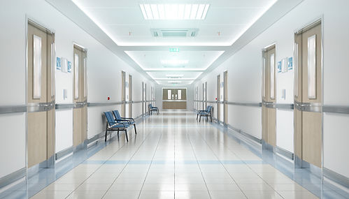 a clean, bright hospital corridor. The hallway is long with a shiny tiled floor and white walls. Doors to patient rooms line both sides, each with a small window and identification signs. There are several blue chairs spaced along the walls for waiting. The ceiling has recessed lighting and an illuminated emergency exit sign towards the corridor end. The overall atmosphere is sterile and orderly, typical of a medical facility.