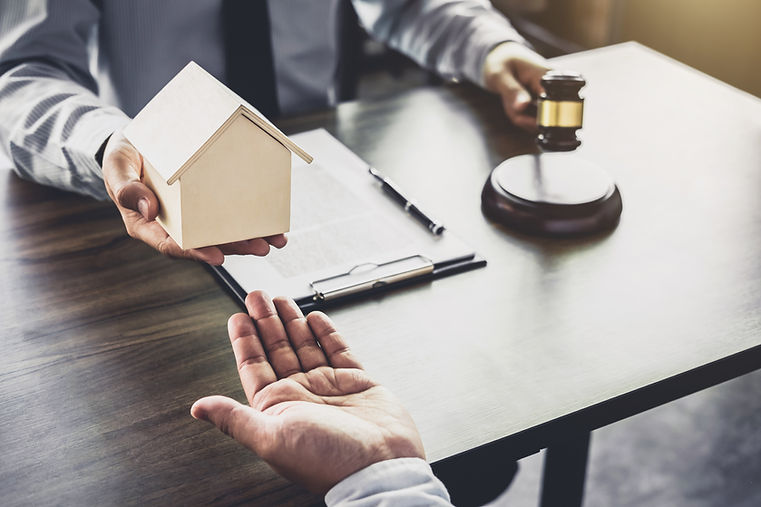 A person in formal attire holds a wooden house model over a desk, handing it to another's outstretched hand