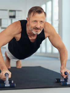Man doing push-ups on handles in a bright room, wearing a black tank top. He's on a black mat, with a focused expression.