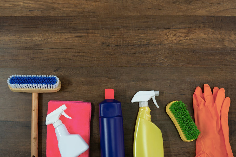 Cleaning supplies arranged on a wooden surface, including a brush, sponges, various spray bottles, and gloves.