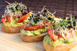 Plant-based avocado toast topped with sliced tomatoes and microgreens on rustic bread, photographed close-up on a wooden board.