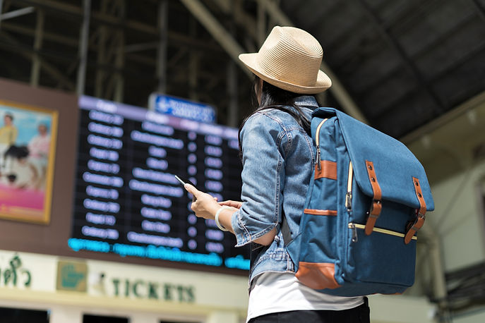 A traveler wearing a hat and blue backpack checks a phone in front of a departure board at a train station