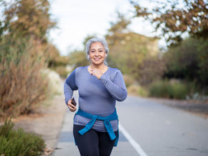 Woman jogging on a tree-lined path, wearing a purple top, black leggings, and blue sweater around waist. She holds a phone and smiles.