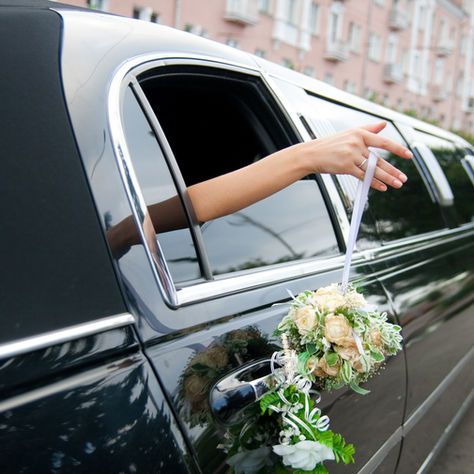 A hand holding a bouquet leans out of a black limousine window, adorned with flowers, on a city street with pink buildings in the background.