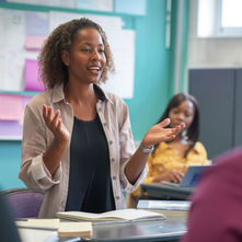 Woman speaks passionately to a group in a classroom, gesturing with hands. Open book on table, bulletin board in background. Classroom setting.