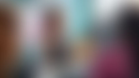 Woman speaks passionately to a group in a classroom, gesturing with hands. Open book on table, bulletin board in background. Classroom setting.