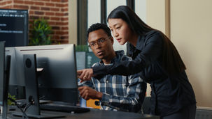 Two people at a computer, with one pointing at the screen. Brick wall and plants in the background. They appear focused and engaged.