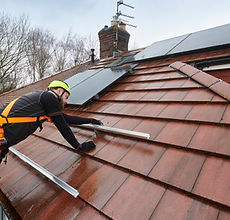 A worker in a safety harness and helmet installs solar panels on a sloped red roof