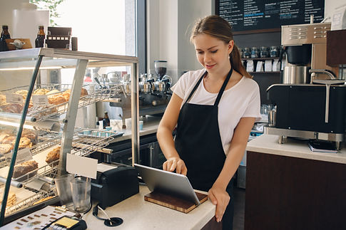 Barista in a black apron using a tablet at a cozy café, smiling. Pastries in a display case and coffee machines in the background.
