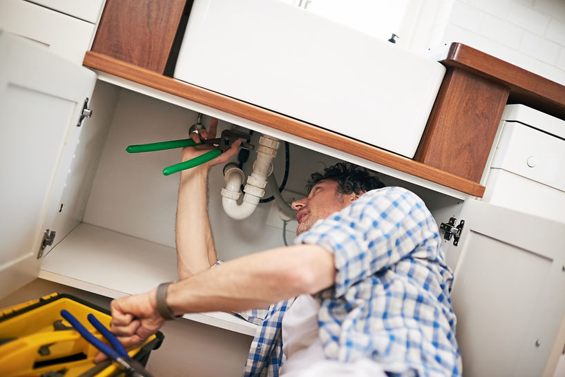 Tradesperson working underneath a wooden cabinet fixture fixing a pipe with tools