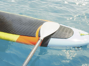 Paddle rests on a colorful paddleboard floating on sparkling blue water. The board has a black textured surface and vibrant yellow and orange sides.