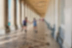 Two people rollerblading through a long colonnade with a patterned tile floor and tall classical columns, enjoying a sunny day in an architectural promenade.